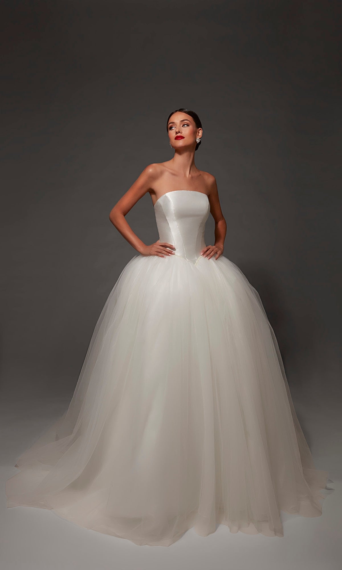 A woman wearing the Alyce 7182 Long Wedding Dress, featuring a strapless mikado bodice and a full, ball gown skirt, stands against a dark background with her hands on her hips.